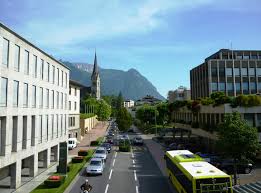 A city street features modern buildings, a bus, a church spire, and distant mountains under a clear blue sky.