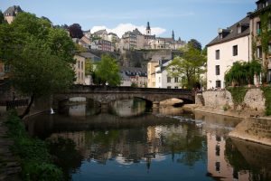 A picturesque European cityscape with traditional buildings, a stone bridge, lush greenery, and a clear sky.