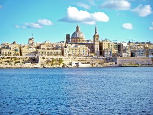 A coastal cityscape under a blue sky with a prominent dome and spires, viewed from across a body of water.