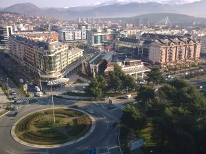 Aerial view of a cityscape with buildings, roads, and a roundabout in the foreground, with mountains in the background under a clear sky.