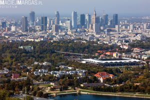 Aerial view of a cityscape with modern skyscrapers in the background and a large park with green spaces and a body of water in the foreground.