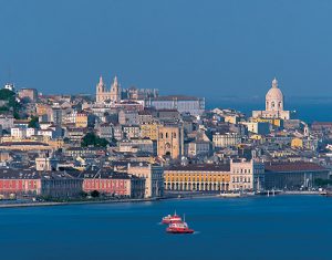 A coastal cityscape during daylight with historic buildings, a prominent church, and a red boat on the water in the foreground.