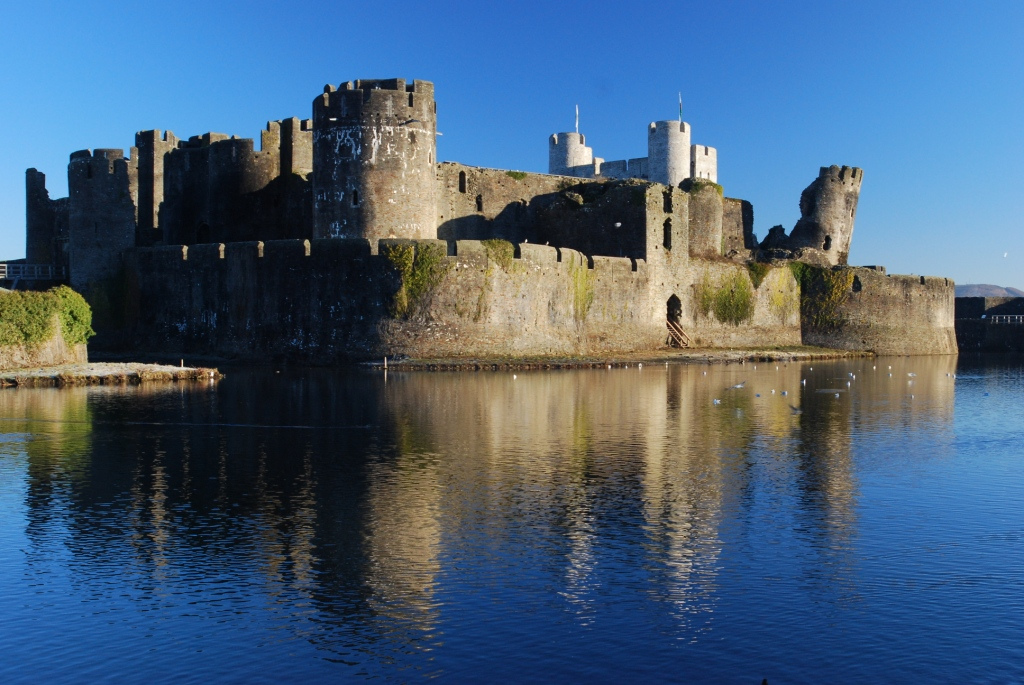 A medieval castle with multiple towers and walls reflected in a calm body of water under a clear blue sky.