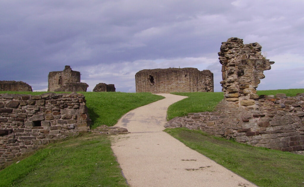 A winding path leading through grassy fields to the ruins of an ancient stone fortress under a cloudy sky.