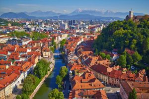 Aerial view of a European city with a central river, surrounded by greenery and mountains under a clear sky.