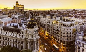 Aerial view of a city at dusk, showcasing illuminated streets and buildings with a warm sunset in the background.