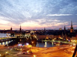 A dusk cityscape with lit buildings and spires, a river, and reflected lights on the water.