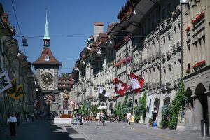 A street view in a European city with traditional architecture, a clock tower, Swiss flags, and people walking.