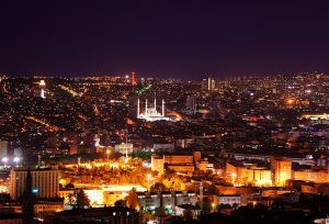 A nighttime cityscape with illuminated buildings and streets, prominently featuring a mosque with minarets lit up in the center.