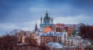 A twilight panorama of a historic city, featuring a church with green domes and golden crosses, amid buildings and bare trees.