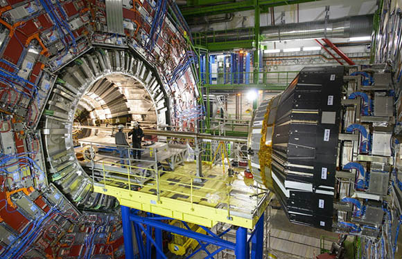 A person stands on a platform inside a large, circular particle detector with complex machinery and colorful wiring visible.