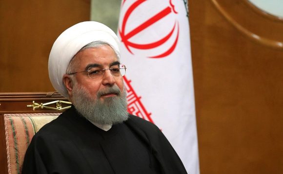 A man with a white beard and glasses wearing a white turban and dark robe sitting in front of an emblem with Persian script.