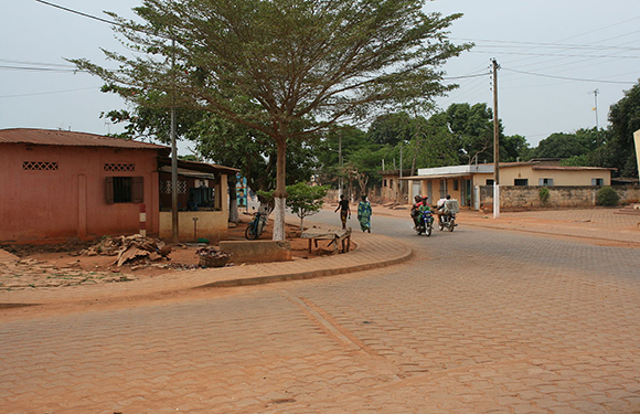 A rural street scene with red dirt roads, people, bicycles, simple buildings, trees, and a cloudy sky.