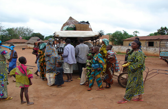 A group of people in colorful traditional clothing gathered around a market area with a thatched structure in the background.