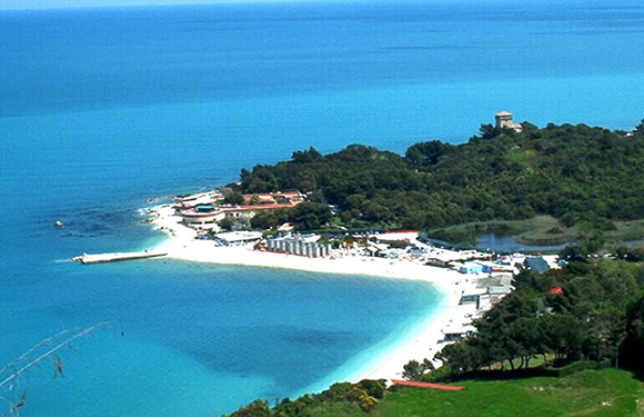 Aerial view of a coastal landscape with a clear blue sea, a curved beachfront, greenery, and buildings.