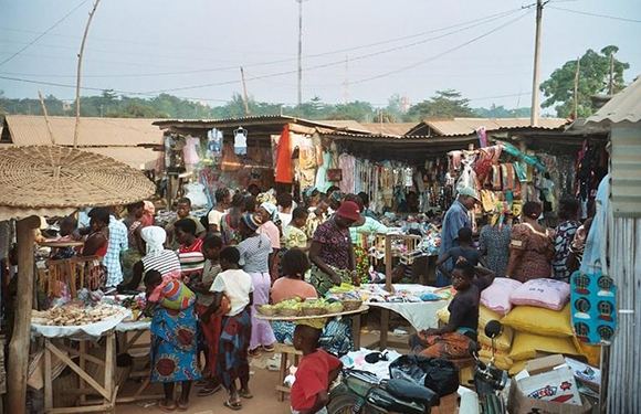 A bustling outdoor market with various stalls displaying clothes and other goods, with numerous people browsing and interacting.