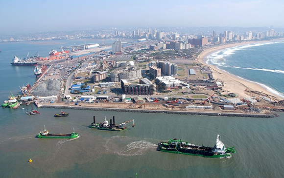 Aerial view of a coastal city with skyscrapers near the beach, a harbor with several ships, and construction areas along the shoreline.