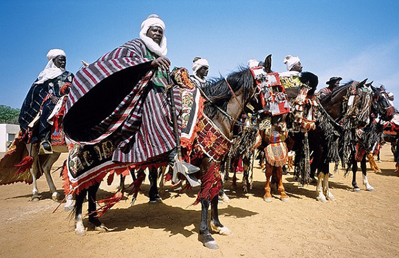 A group of people in traditional attire riding decorated horses at a cultural event under a clear sky.