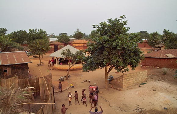 Aerial view of a rural village with thatched and brick houses, a large tree in the center, and several people gathered in open areas.