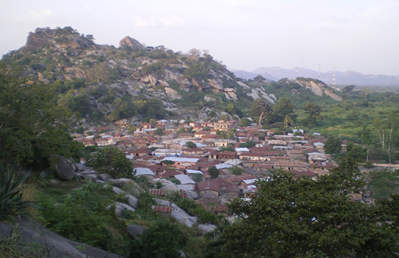 A small town nestled at the base of a rocky hill with dense vegetation, under a hazy sky.