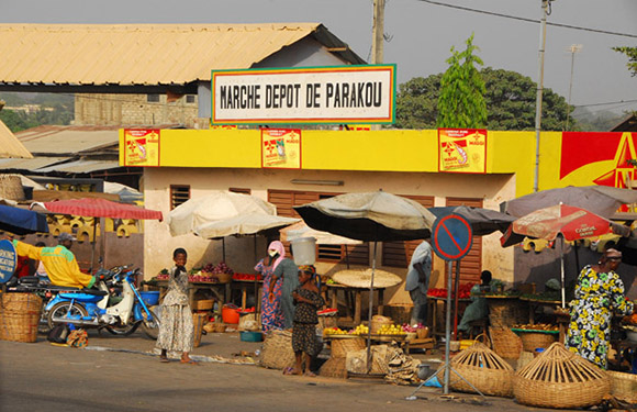 A vibrant street market under colorful umbrellas in a tropical setting, with a sign "MARCHE DEPOT DE PARAKOU" on a colorful building.