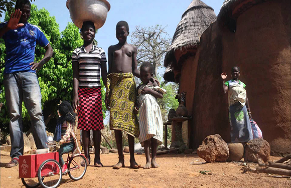 People stand by mud houses with thatched roofs, some carrying items on their heads, with a red bicycle in the foreground.