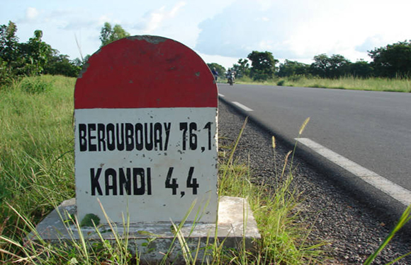 A road sign shows distances to Beroubouay and Kandi, set against a backdrop of greenery and a partly cloudy sky.