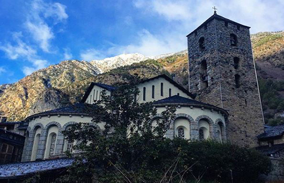 A stone church with a tall bell tower in the foreground, set against a backdrop of tree-covered mountains under a partly cloudy sky.