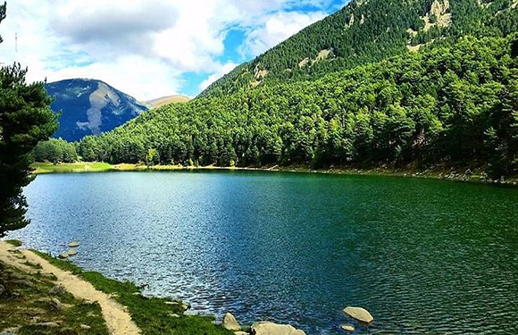 A scenic view of a tranquil lake surrounded by lush green trees with mountains in the background under a partly cloudy sky.