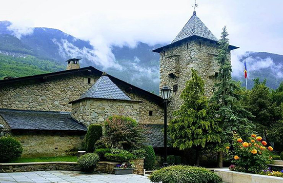 A picturesque view of a historic stone building with a tower, surrounded by lush greenery and mountains in the background, under an overcast sky.