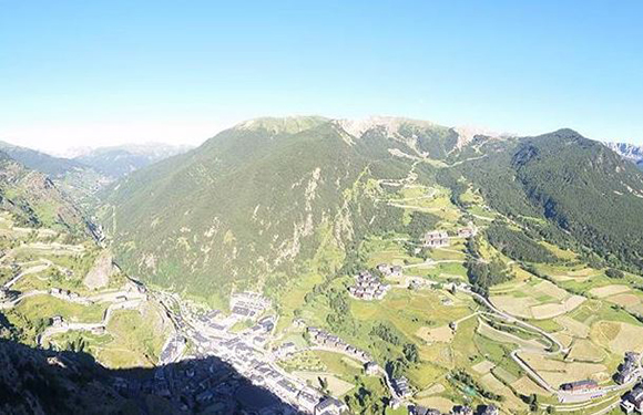 Aerial view of a mountainous landscape with green slopes, some patches of forest, and a small village with buildings clustered in the valley.