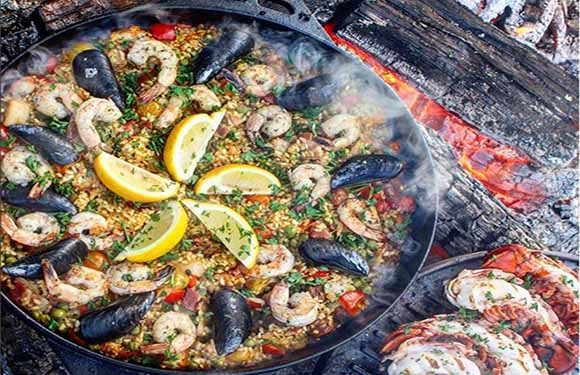 A colorful seafood paella in a large pan with lemon slices, mussels, and shrimp, cooked over an open flame.