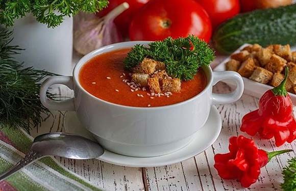 A bowl of tomato soup with croutons and a garnish, surrounded by fresh tomatoes and herbs on a wooden surface.