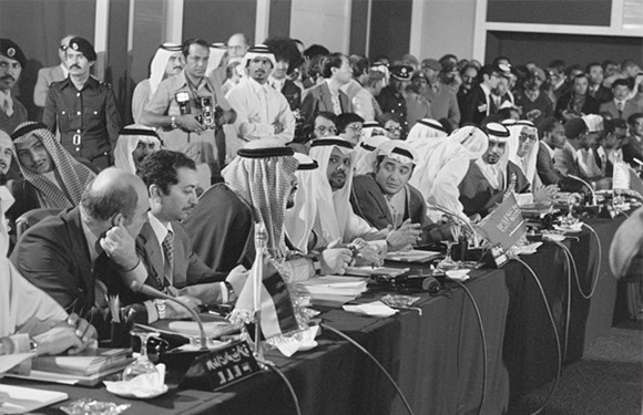 A black and white photo shows men in Middle Eastern and Western attire at a conference, with flags and observers present.