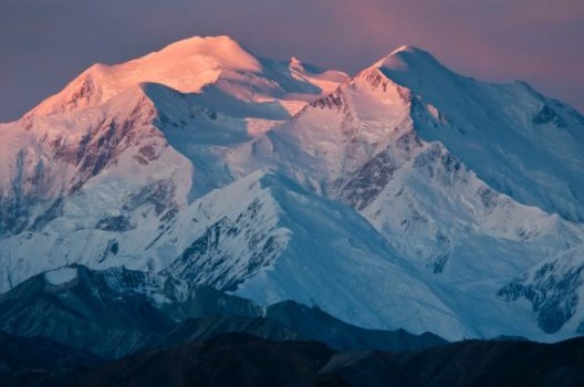 A photograph of snow-covered mountain peaks bathed in the warm glow of a sunrise or sunset.