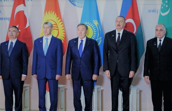 A group of six men in suits standing side by side with flags in the background.