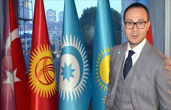 A man in a suit and glasses standing in front of three vertical flags, with a window showing a cityscape in the background.