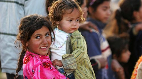 A girl in a pink scarf and necklace carries a child on her back in a crowded place.