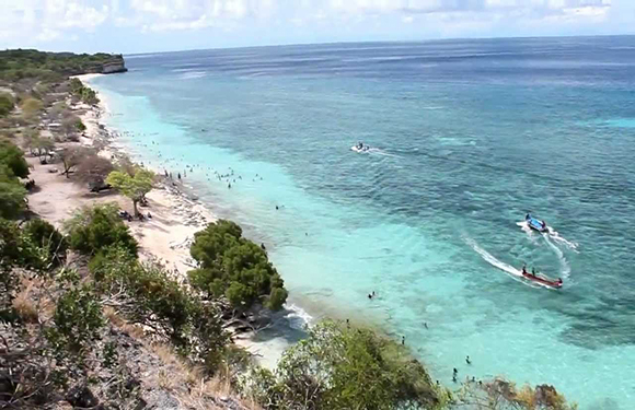 A coastal landscape with clear turquoise waters, several small boats near the shore, and a rocky coastline with sparse vegetation.