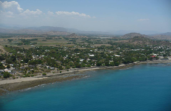 Aerial view of a coastal area with clear blue waters, a beach, clusters of buildings, and a backdrop of rolling hills and greenery.