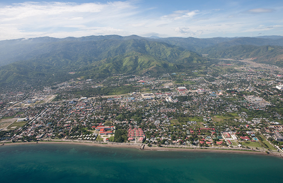 Aerial view of a coastal city near a large body of water, with buildings along the shore and mountains in the background.