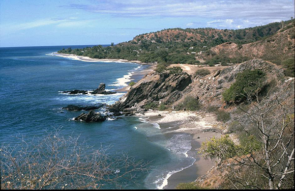 A coastal landscape with a rocky shoreline and sandy beaches, blue ocean waters, and a hilly terrain with sparse vegetation under a clear sky.