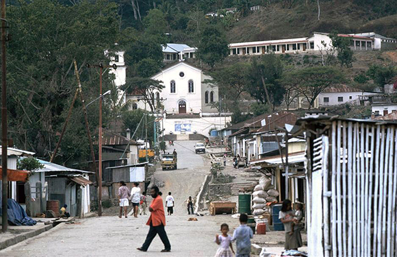 A street view in a small town with a white church at the end of the street, people walking, and buildings on either side.