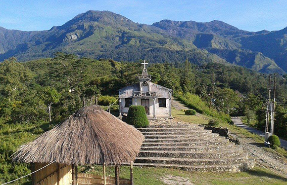 A small church with a cross, at a green mountain base, has steps leading up to it and a thatched-roof structure nearby.