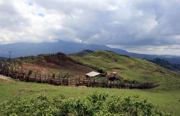 A rural landscape with rolling hills and a small wooden structure surrounded by a fence. Cloudy skies and distant mountains in the background.