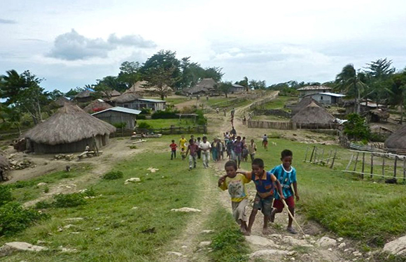 A group of children walking along a path in a rural village with thatched-roof houses and lush greenery in the background.