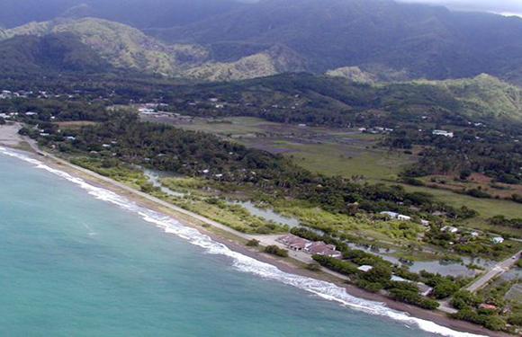 Aerial view of a coastal area with a beach adjacent to a body of water on the left, a line of trees, and a mountainous landscape in the background.