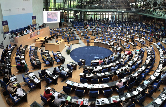 A panoramic view of a large, semi-circular parliamentary chamber with rows of desks, seated individuals, and a central speaking area.