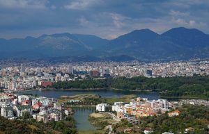 A cityscape with buildings, a large water body in front, mountains behind, under a cloudy sky.