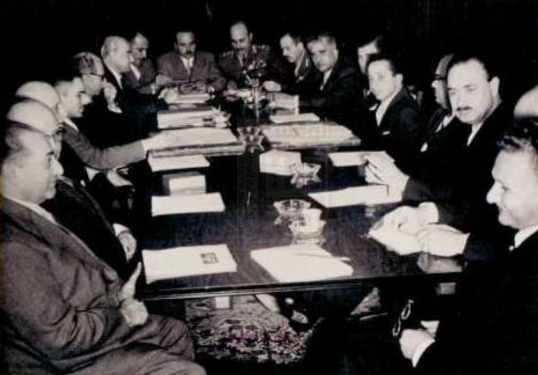 A black and white historical photo of a group of men in suits sitting around a long table during a meeting.